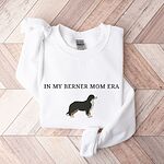 White crewneck sweatshirt laid flat on herringbone wood, embroidered with In My Berner Mom Era above a standing Bernese mountain dog