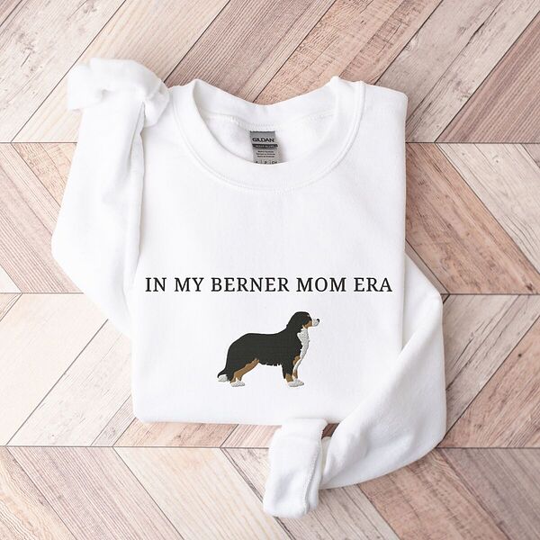 White crewneck sweatshirt laid flat on herringbone wood, embroidered with In My Berner Mom Era above a standing Bernese mountain dog
