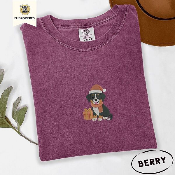 Berry Comfort Colors T-shirt embroidered with a tricolor Bernese mountain dog puppy in a Santa hat and knit scarf sitting beside a wrapped gift