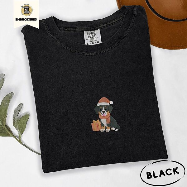 Black Comfort Colors T-shirt embroidered with a tricolor Bernese mountain dog puppy in a Santa hat and knit scarf sitting beside a wrapped gift