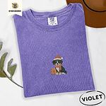 Violet Comfort Colors T-shirt embroidered with a tricolor Bernese mountain dog puppy in a Santa hat and knit scarf sitting beside a wrapped gift