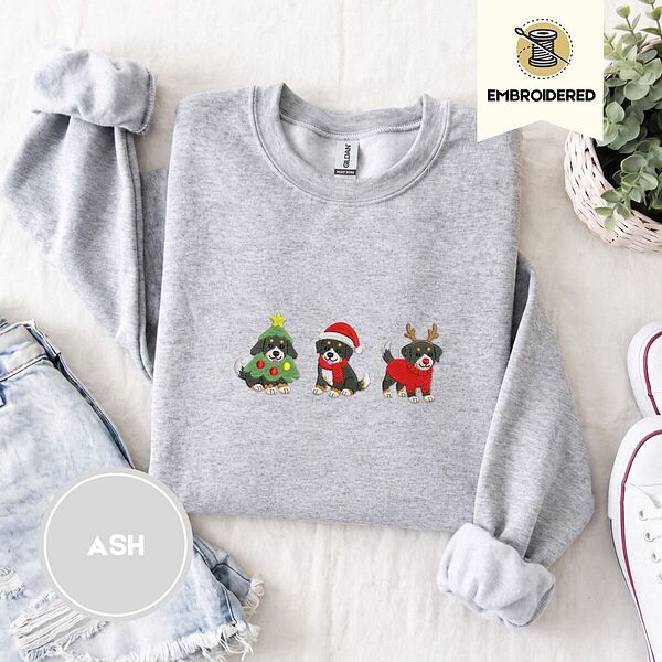 Ash crewneck sweatshirt folded with denim shorts, white sneakers and a potted plant, embroidered with three Bernese mountain dog puppies in a Christmas tree costume, a Santa hat with a red scarf and reindeer antlers with a red sweater