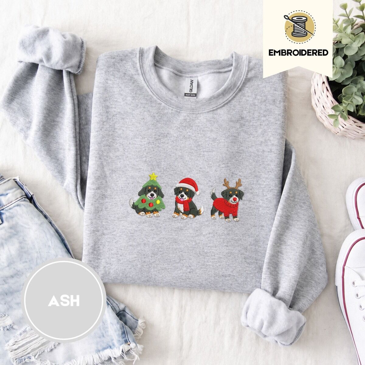 Bernese Mountain Dog Christmas Sweatshirt – Ash Ash crewneck sweatshirt folded with denim shorts, white sneakers and a potted plant, embroidered with three Bernese mountain dog puppies in a Christmas tree costume, a Santa hat with a red scarf and reindeer antlers with a red sweater