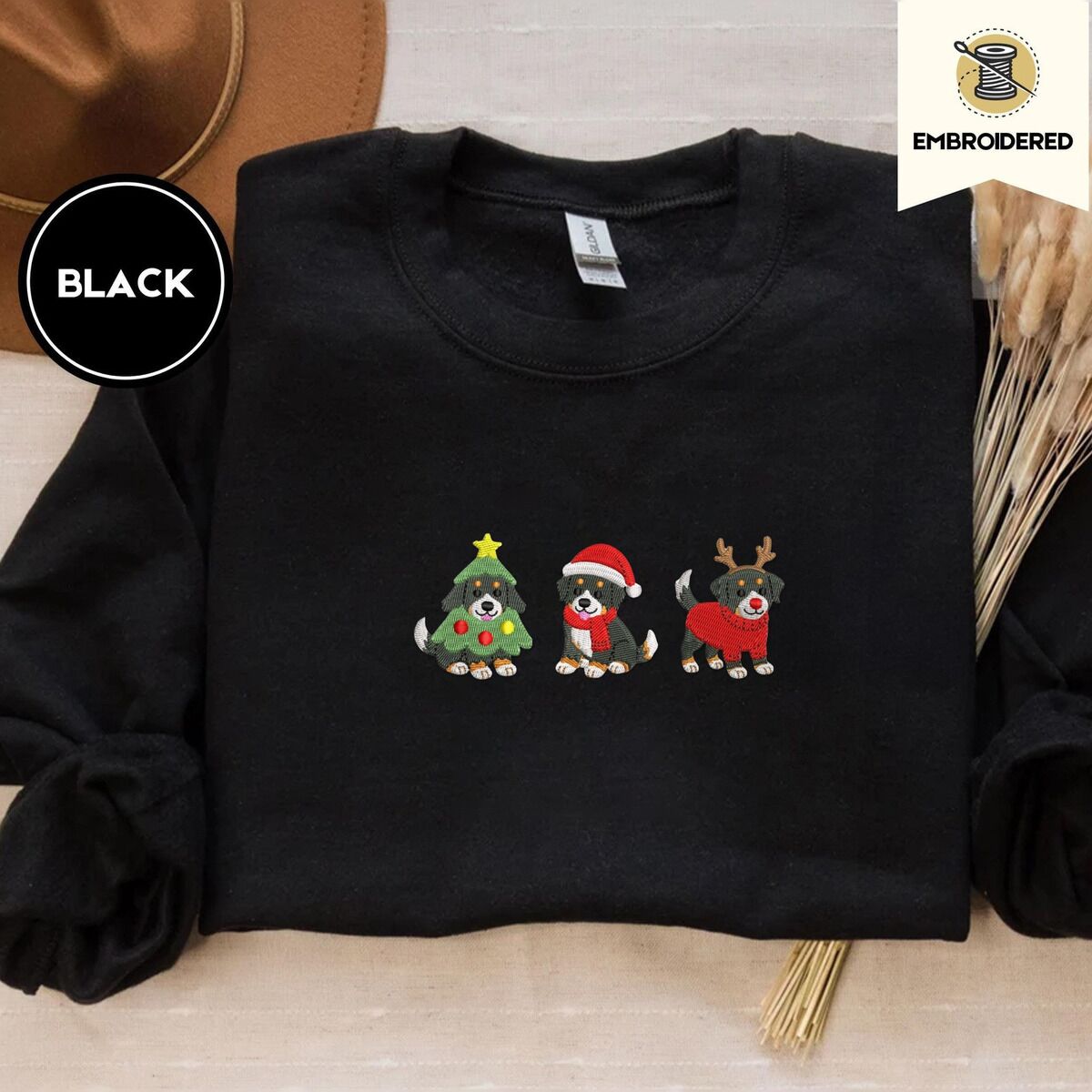 Bernese Mountain Dog Christmas Sweatshirt – Black Black crewneck sweatshirt with a brown felt hat and dried bunny tail grass, embroidered with three Bernese mountain dog puppies in a Christmas tree costume, a Santa hat with a red scarf and reindeer antlers with a red sweater