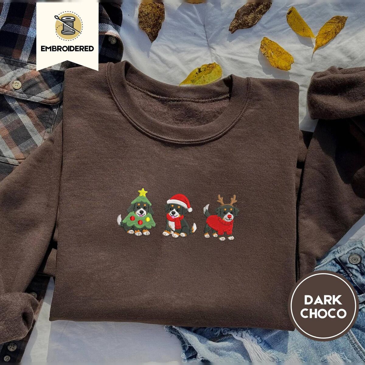 Bernese Mountain Dog Christmas Sweatshirt – Dark Chocolate Dark Chocolate crewneck sweatshirt on a plaid flannel shirt with yellow autumn leaves and denim shorts, embroidered with three Bernese mountain dog puppies in a Christmas tree costume, a Santa hat with a red scarf and reindeer antlers with a red sweater
