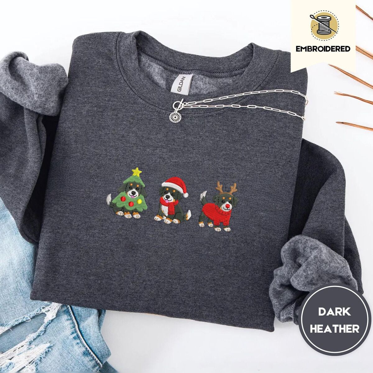 Bernese Mountain Dog Christmas Sweatshirt – Dark Heather Dark Heather crewneck sweatshirt folded with ripped jeans and a silver chain necklace, embroidered with three Bernese mountain dog puppies in a Christmas tree costume, a Santa hat with a red scarf and reindeer antlers with a red sweater