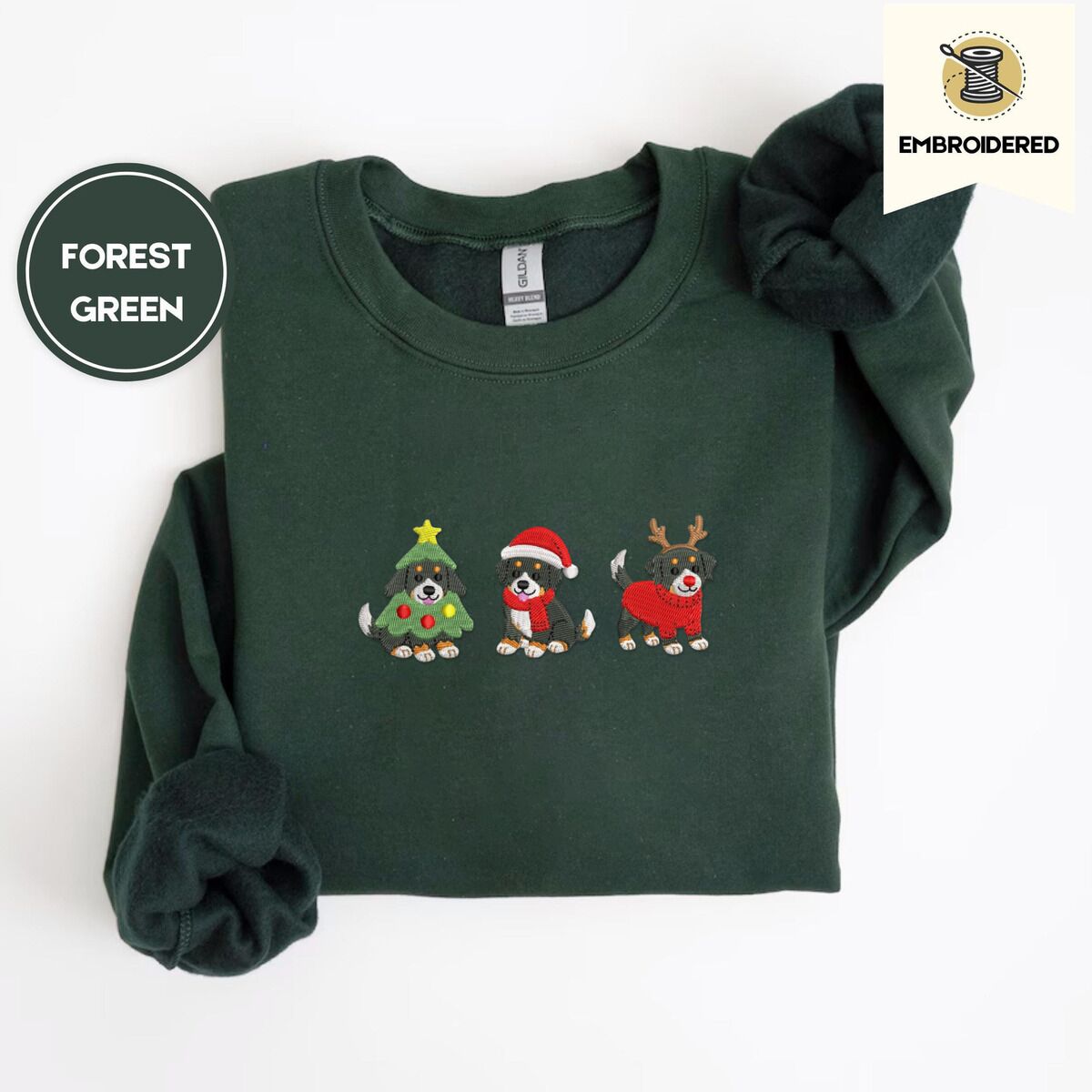 Bernese Mountain Dog Christmas Sweatshirt – Forest Green Forest Green crewneck sweatshirt folded on a white background, embroidered with three Bernese mountain dog puppies in a Christmas tree costume, a Santa hat with a red scarf and reindeer antlers with a red sweater