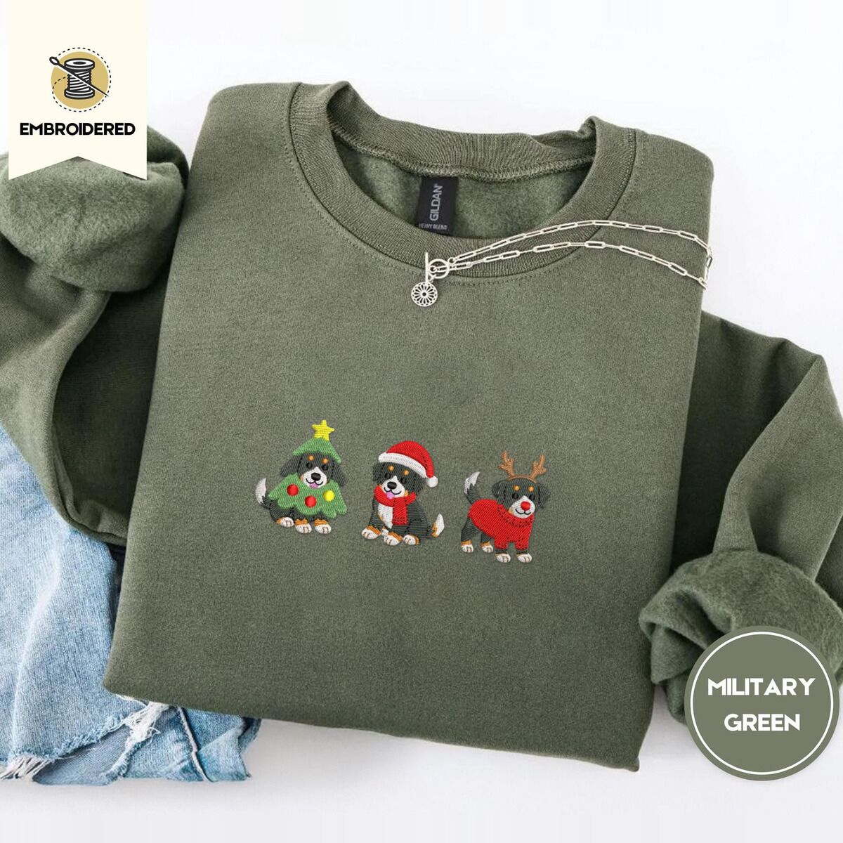 Bernese Mountain Dog Christmas Sweatshirt – Military Green Military Green crewneck sweatshirt folded with light jeans and a silver chain necklace, embroidered with three Bernese mountain dog puppies in a Christmas tree costume, a Santa hat with a red scarf and reindeer antlers with a red sweater