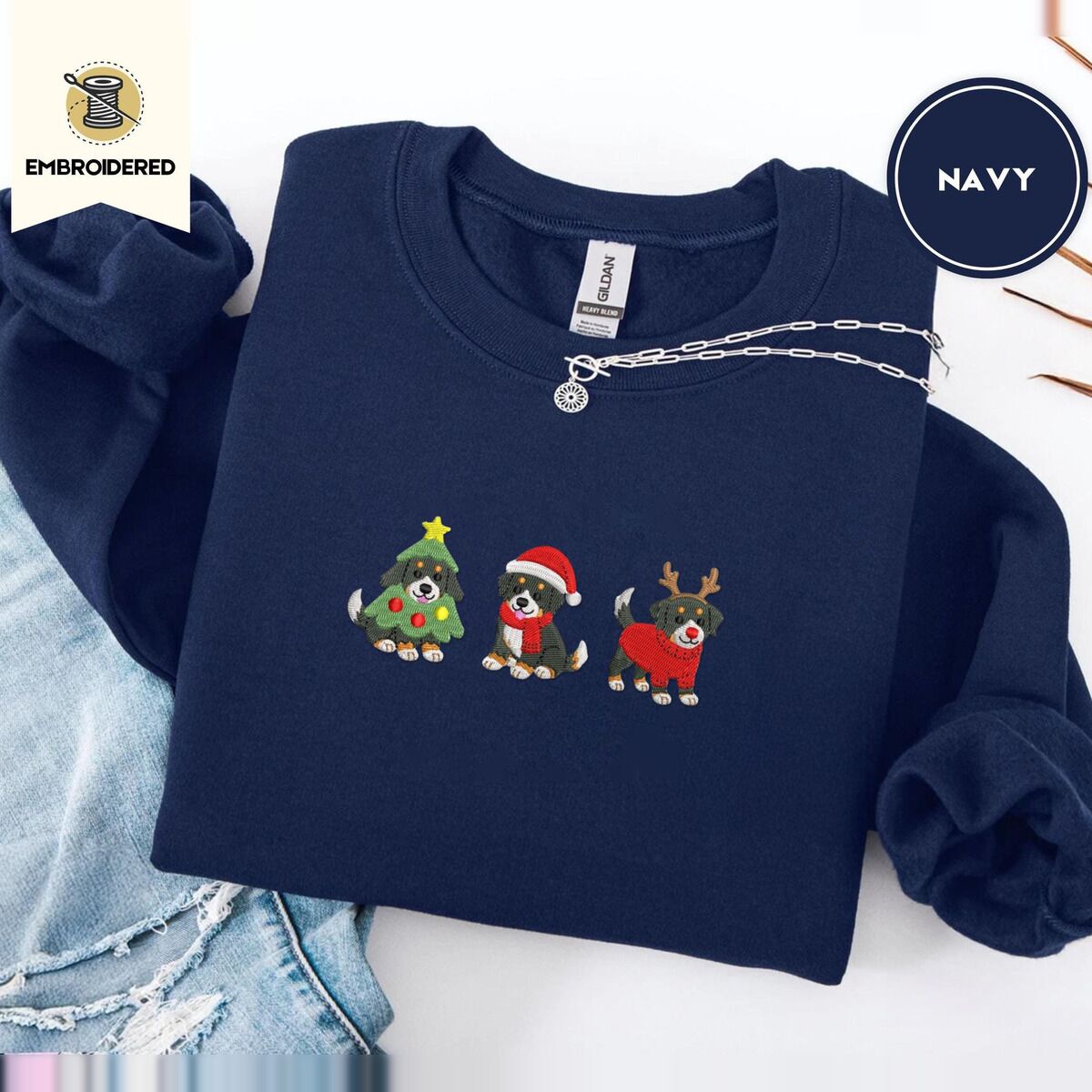 Bernese Mountain Dog Christmas Sweatshirt – Navy Navy crewneck sweatshirt folded with ripped jeans and a silver chain necklace, embroidered with three Bernese mountain dog puppies in a Christmas tree costume, a Santa hat with a red scarf and reindeer antlers with a red sweater