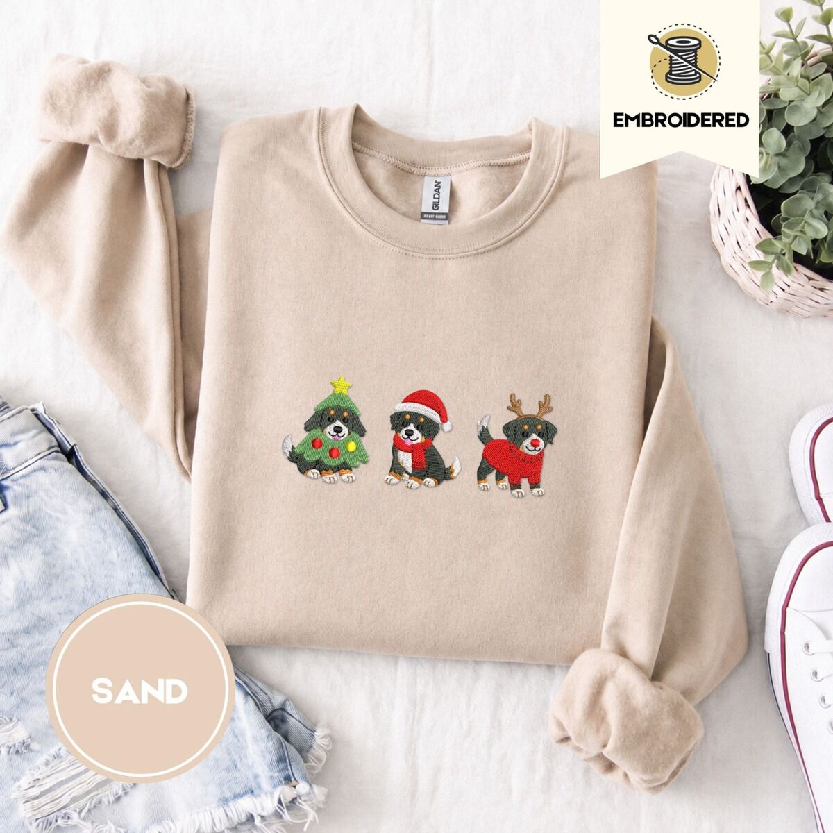 Bernese Mountain Dog Christmas Sweatshirt – Sand Sand crewneck sweatshirt folded with denim shorts, white sneakers and a potted plant, embroidered with three Bernese mountain dog puppies in a Christmas tree costume, a Santa hat with a red scarf and reindeer antlers with a red sweater