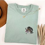 Bay T-shirt laid flat with a necklace and dried palm leaves, embroidered with a tricolor Berner head in profile on the left chest