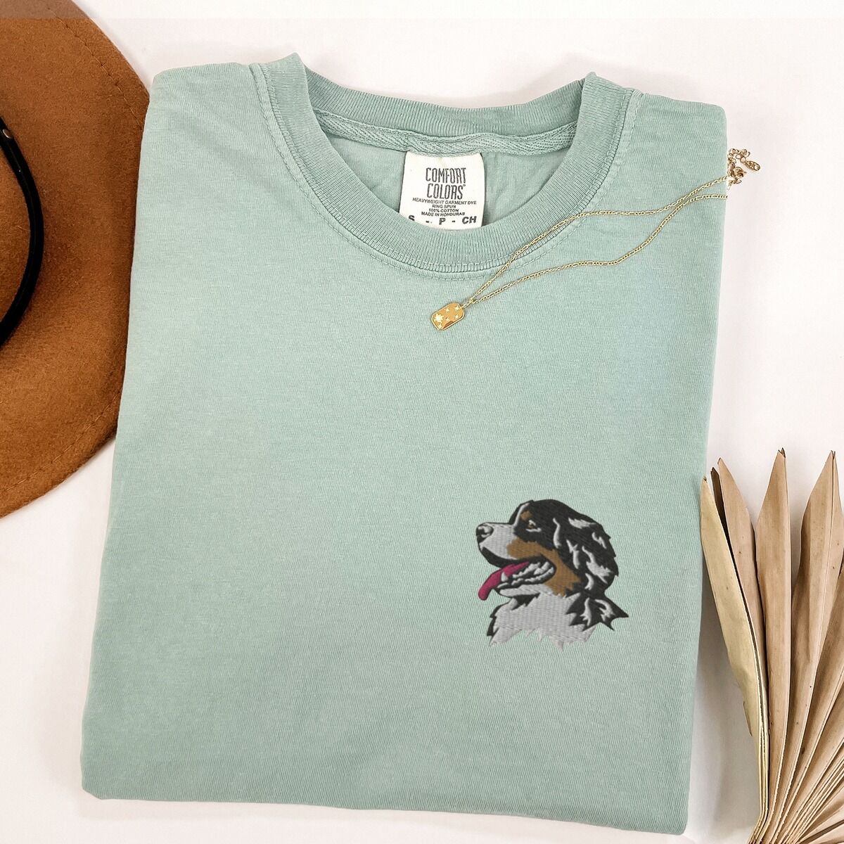 Bernese Mountain Dog Head Shirt – Bay Bay T-shirt laid flat with a necklace and dried palm leaves, embroidered with a tricolor Berner head in profile on the left chest