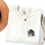 Ivory T-shirt laid flat with a felt hat and a necklace, embroidered with a shaggy Bernese mountain dog head on the left chest