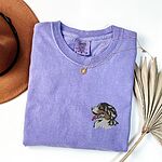Violet T-shirt laid flat with a felt hat and dried palm leaves, embroidered with a tricolor Berner head with its tongue out on the left chest