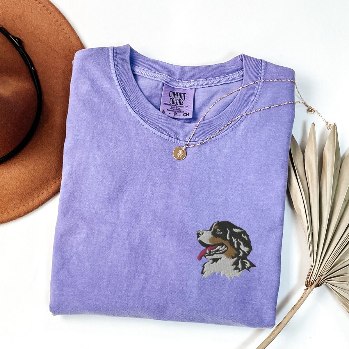 Bernese Mountain Dog Head Shirt – Violet Violet T-shirt laid flat with a felt hat and dried palm leaves, embroidered with a tricolor Berner head with its tongue out on the left chest