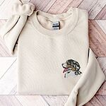 Sand crewneck sweatshirt laid flat on herringbone wood, embroidered with a Bernese mountain dog head in profile with its pink tongue out on the left chest