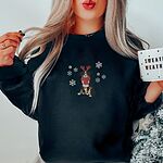 Woman holding a mug in a Black crewneck sweatshirt embroidered with a sitting tricolor Bernese mountain dog in red reindeer antlers and a red knit scarf, framed by snowflakes