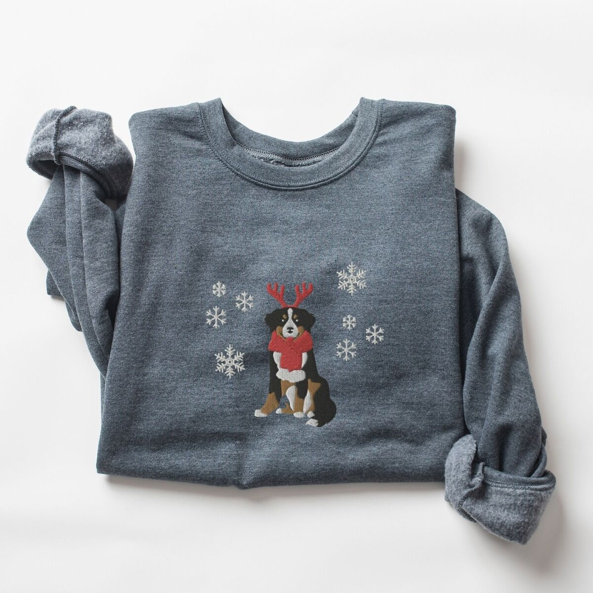 Bernese Mountain Dog Reindeer Sweatshirt – Dark Heather Dark Heather crewneck sweatshirt laid flat with rolled sleeves, embroidered with a sitting tricolor Bernese mountain dog in red reindeer antlers and a red knit scarf, framed by snowflakes