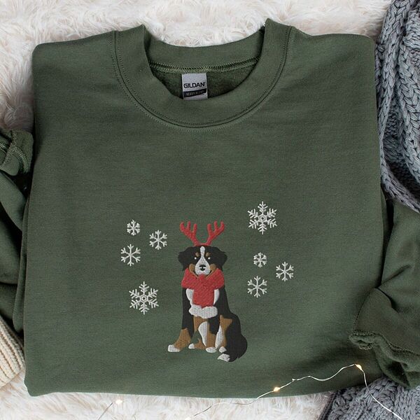 Military Green crewneck sweatshirt laid flat on a faux fur throw, embroidered with a sitting tricolor Bernese mountain dog in red reindeer antlers and a red knit scarf, framed by snowflakes