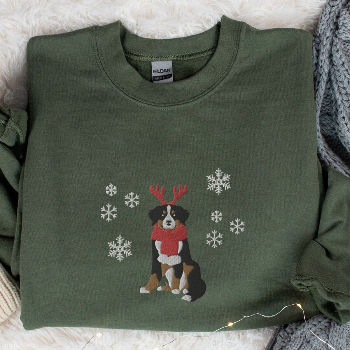 Bernese Mountain Dog Reindeer Sweatshirt – Military Green Military Green crewneck sweatshirt laid flat on a faux fur throw, embroidered with a sitting tricolor Bernese mountain dog in red reindeer antlers and a red knit scarf, framed by snowflakes