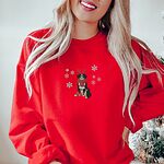 Woman by a snowy Christmas tree in a Red crewneck sweatshirt embroidered with a sitting tricolor Bernese mountain dog in red reindeer antlers and a red knit scarf, framed by snowflakes