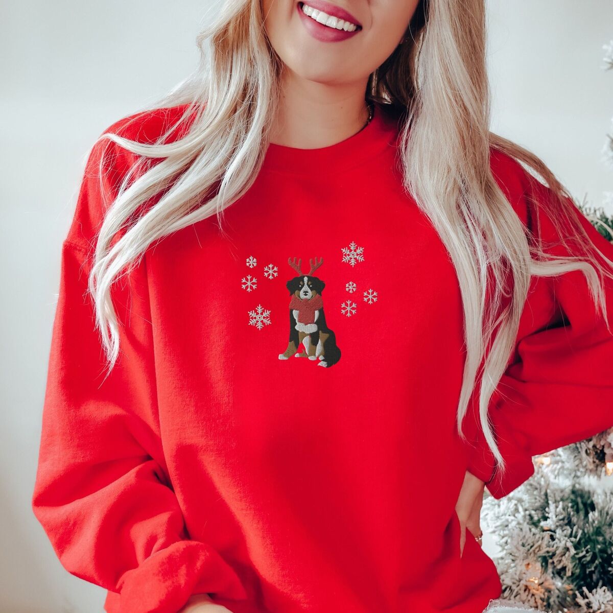 Bernese Mountain Dog Reindeer Sweatshirt - Red Woman by a snowy Christmas tree in a Red crewneck sweatshirt embroidered with a sitting tricolor Bernese mountain dog in red reindeer antlers and a red knit scarf, framed by snowflakes