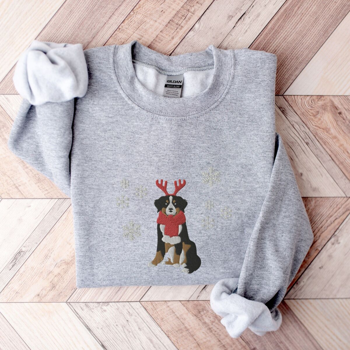 Bernese Mountain Dog Reindeer Sweatshirt - Sport Grey Sport Grey crewneck sweatshirt laid flat on a chevron wood floor, embroidered with a sitting tricolor Bernese mountain dog in red reindeer antlers and a red knit scarf, framed by snowflakes