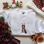 White crewneck sweatshirt laid flat with a plaid scarf, pine and a wooden XMAS sign, embroidered with a sitting tricolor Bernese mountain dog in red reindeer antlers and a red knit scarf, framed by snowflakes