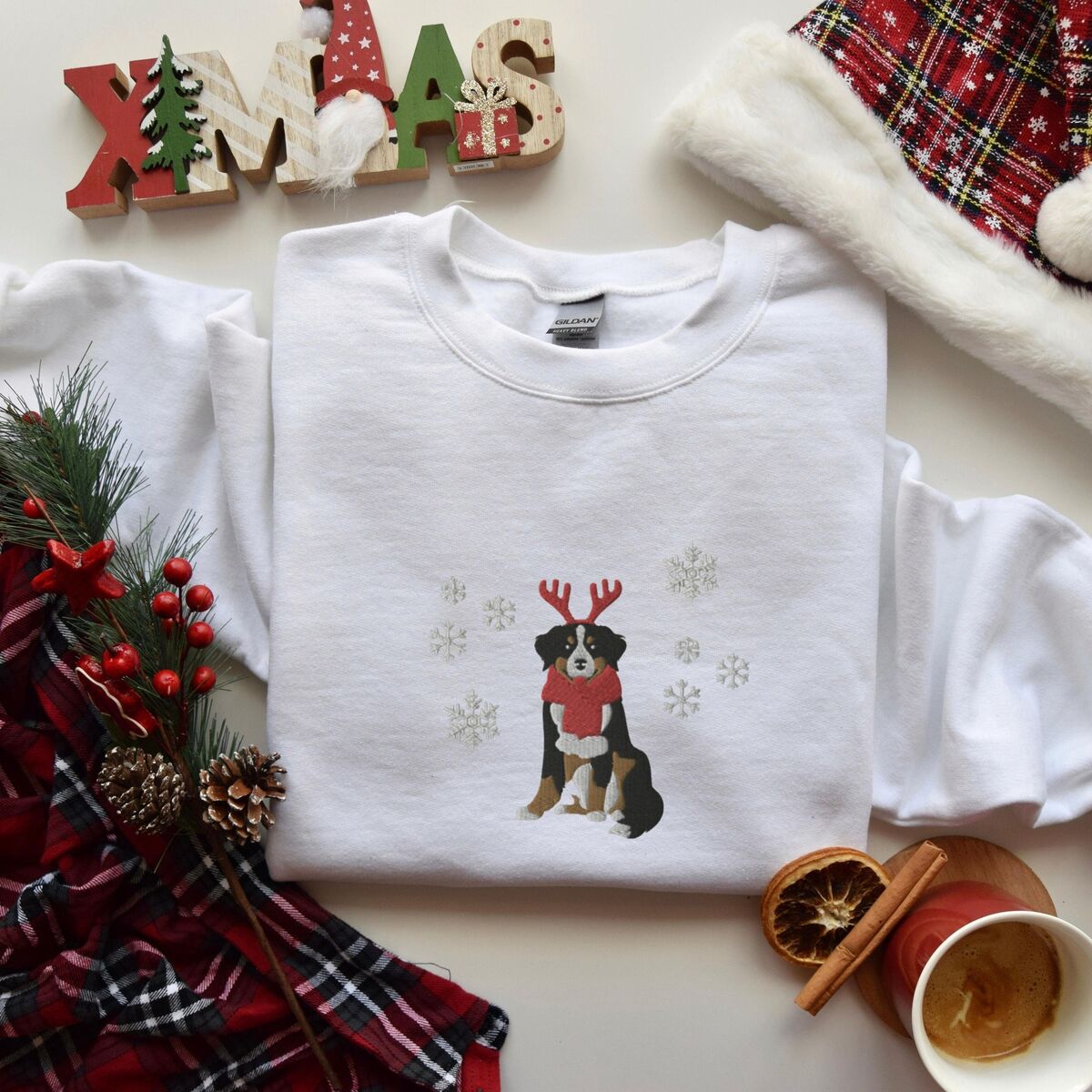 Bernese Mountain Dog Reindeer Sweatshirt - White White crewneck sweatshirt laid flat with a plaid scarf, pine and a wooden XMAS sign, embroidered with a sitting tricolor Bernese mountain dog in red reindeer antlers and a red knit scarf, framed by snowflakes