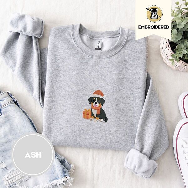 Ash crewneck sweatshirt folded with denim shorts, white sneakers and a potted plant, embroidered with a tricolor Bernese mountain dog puppy in a Santa hat and knit scarf sitting beside a wrapped gift