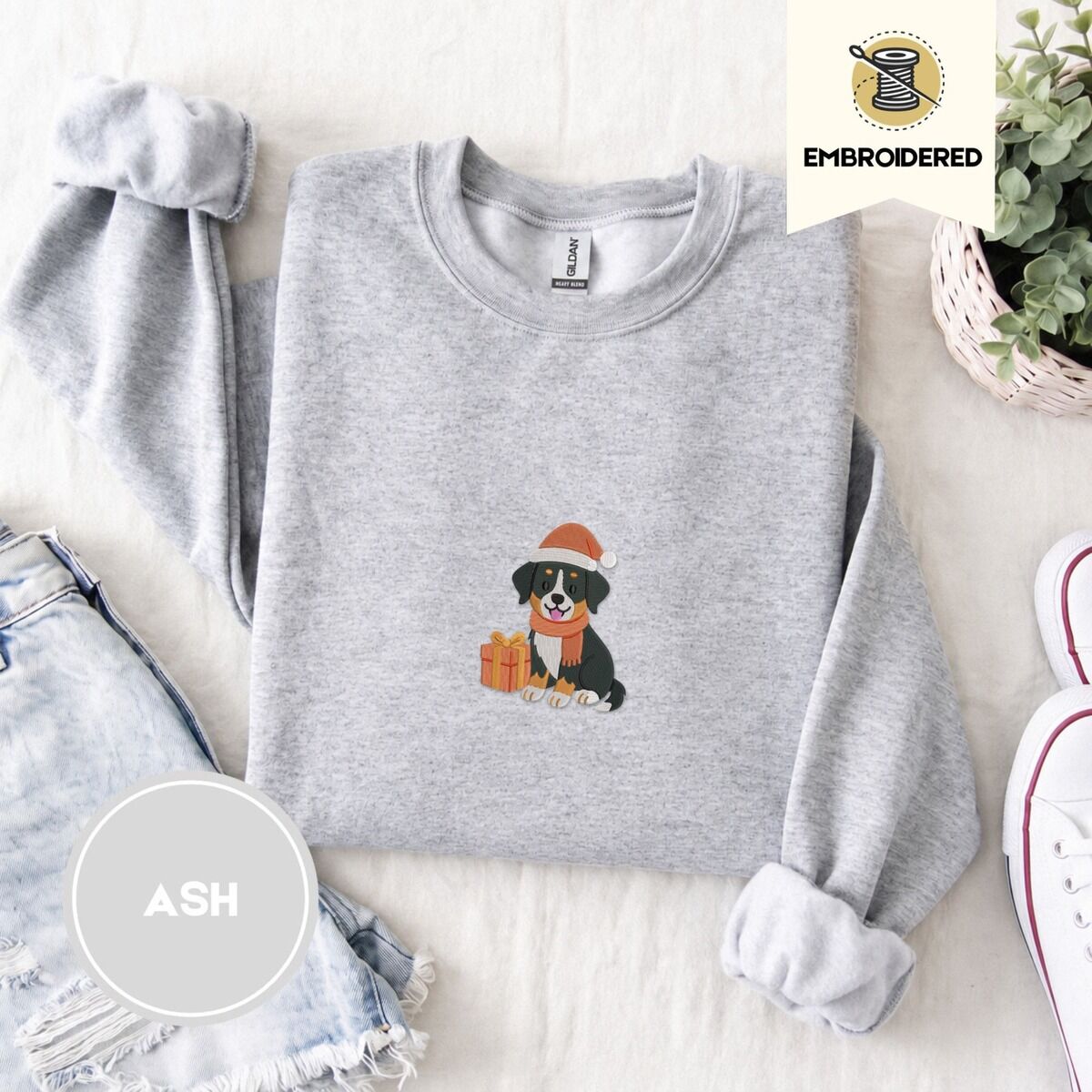 Bernese Mountain Dog Santa Hat Sweatshirt – Ash Ash crewneck sweatshirt folded with denim shorts, white sneakers and a potted plant, embroidered with a tricolor Bernese mountain dog puppy in a Santa hat and knit scarf sitting beside a wrapped gift