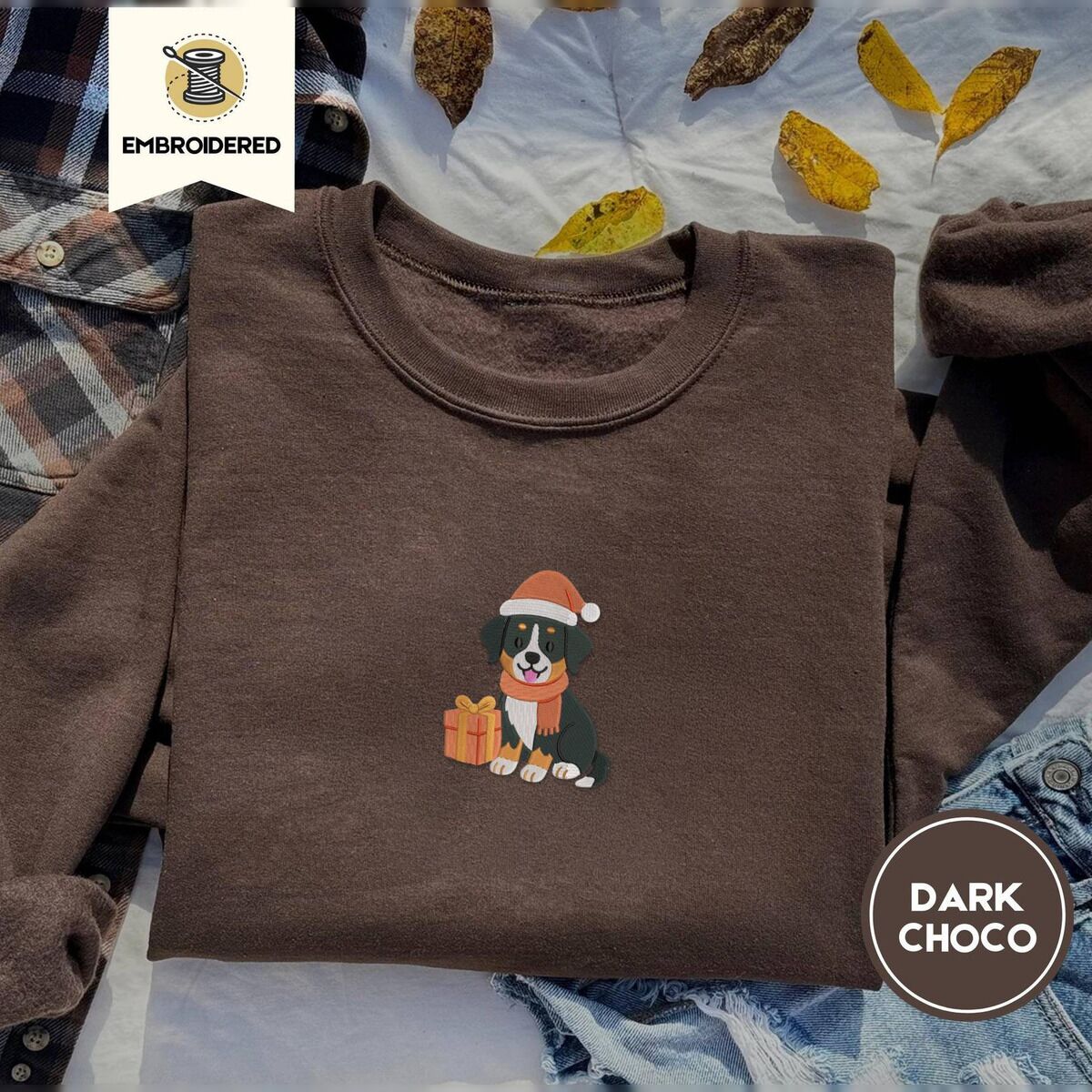 Bernese Mountain Dog Santa Hat Sweatshirt – Dark Chocolate Dark Chocolate crewneck sweatshirt on a plaid flannel shirt with yellow autumn leaves and denim shorts, embroidered with a tricolor Bernese mountain dog puppy in a Santa hat and knit scarf sitting beside a wrapped gift