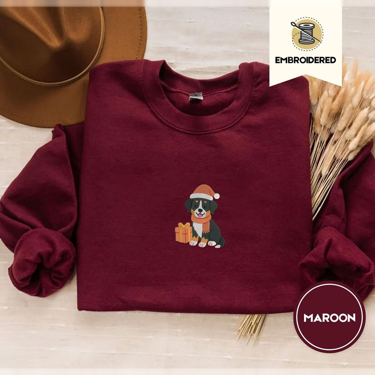 Bernese Mountain Dog Santa Hat Sweatshirt – Maroon Maroon crewneck sweatshirt with a brown felt hat and dried bunny tail grass, embroidered with a tricolor Bernese mountain dog puppy in a Santa hat and knit scarf sitting beside a wrapped gift