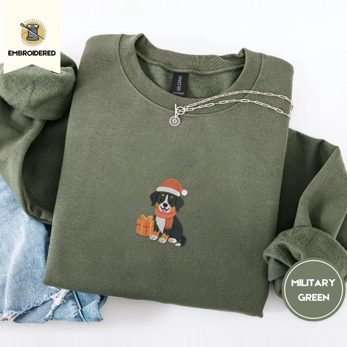 Bernese Mountain Dog Santa Hat Sweatshirt – Military Green Military Green crewneck sweatshirt folded with light jeans and a silver chain necklace, embroidered with a tricolor Bernese mountain dog puppy in a Santa hat and knit scarf sitting beside a wrapped gift