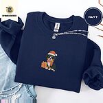 Navy crewneck sweatshirt folded with ripped jeans and a silver chain necklace, embroidered with a tricolor Bernese mountain dog puppy in a Santa hat and knit scarf sitting beside a wrapped gift