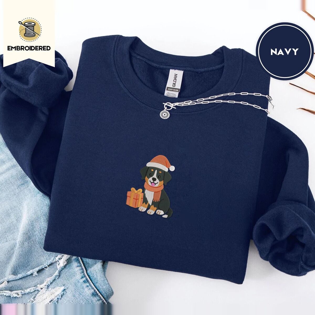 Bernese Mountain Dog Santa Hat Sweatshirt – Navy Navy crewneck sweatshirt folded with ripped jeans and a silver chain necklace, embroidered with a tricolor Bernese mountain dog puppy in a Santa hat and knit scarf sitting beside a wrapped gift