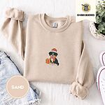 Sand crewneck sweatshirt folded with denim shorts, white sneakers and a potted plant, embroidered with a tricolor Bernese mountain dog puppy in a Santa hat and knit scarf sitting beside a wrapped gift
