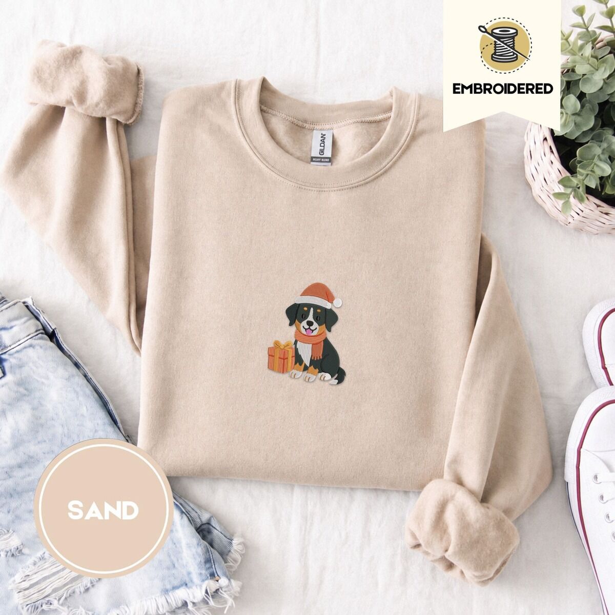 Bernese Mountain Dog Santa Hat Sweatshirt – Sand Sand crewneck sweatshirt folded with denim shorts, white sneakers and a potted plant, embroidered with a tricolor Bernese mountain dog puppy in a Santa hat and knit scarf sitting beside a wrapped gift