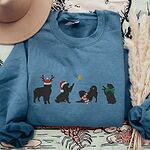 Indigo Blue crewneck sweatshirt laid flat with a hat and dried pampas, embroidered with four black Bernese mountain dog silhouettes in antlers, a Santa hat, a striped scarf and a green bow under a gold star
