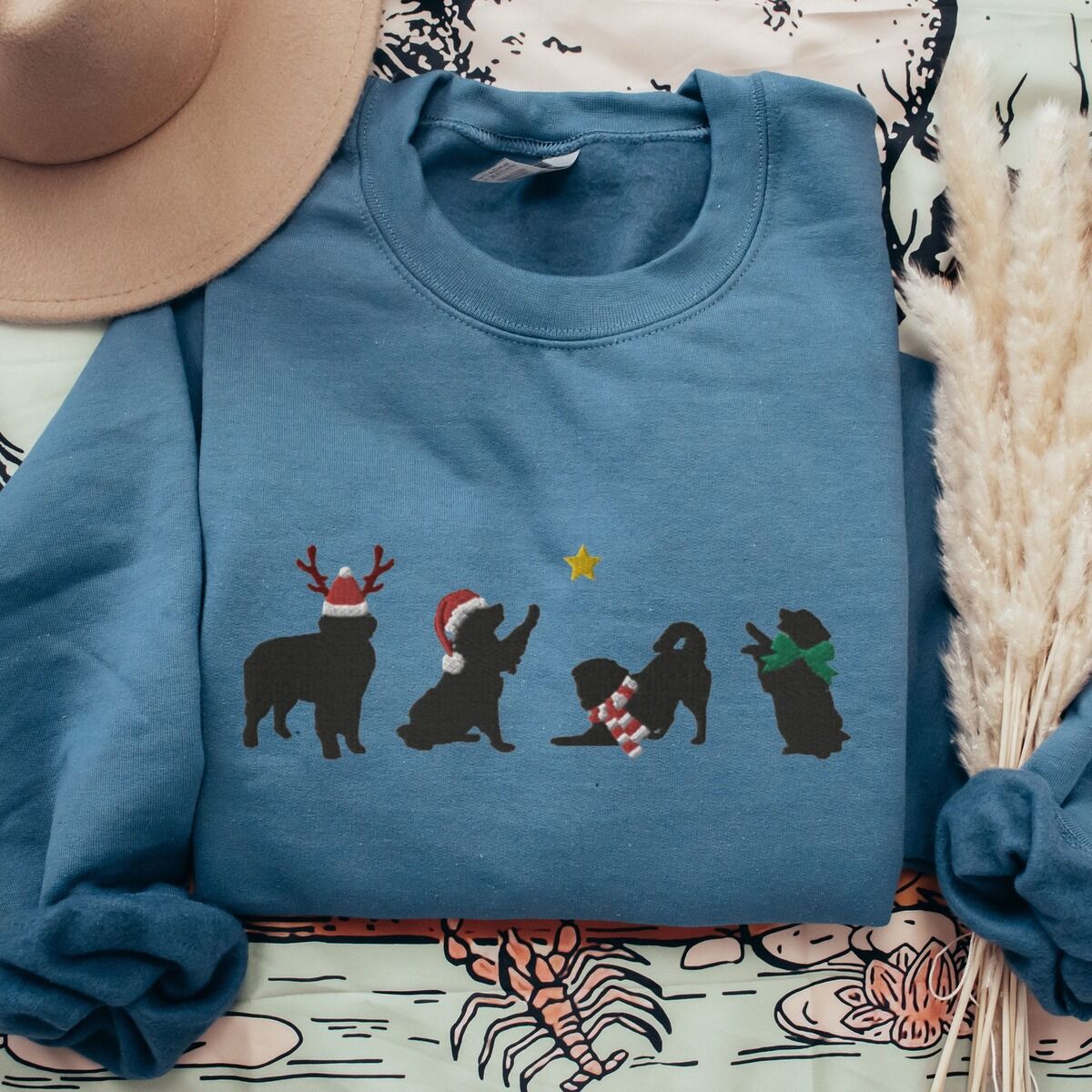 Bernese Mountain Dog Silhouette Sweatshirt – Indigo Blue Indigo Blue crewneck sweatshirt laid flat with a hat and dried pampas, embroidered with four black Bernese mountain dog silhouettes in antlers, a Santa hat, a striped scarf and a green bow under a gold star
