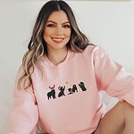 Smiling woman sitting in a Light Pink crewneck sweatshirt embroidered with four black Bernese mountain dog silhouettes in antlers, a Santa hat, a striped scarf and a green bow under a gold star
