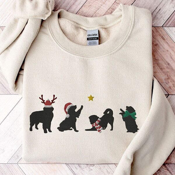 Sand crewneck sweatshirt laid flat on a chevron wood floor, embroidered with four black Bernese mountain dog silhouettes in antlers, a Santa hat, a striped scarf and a green bow under a gold star