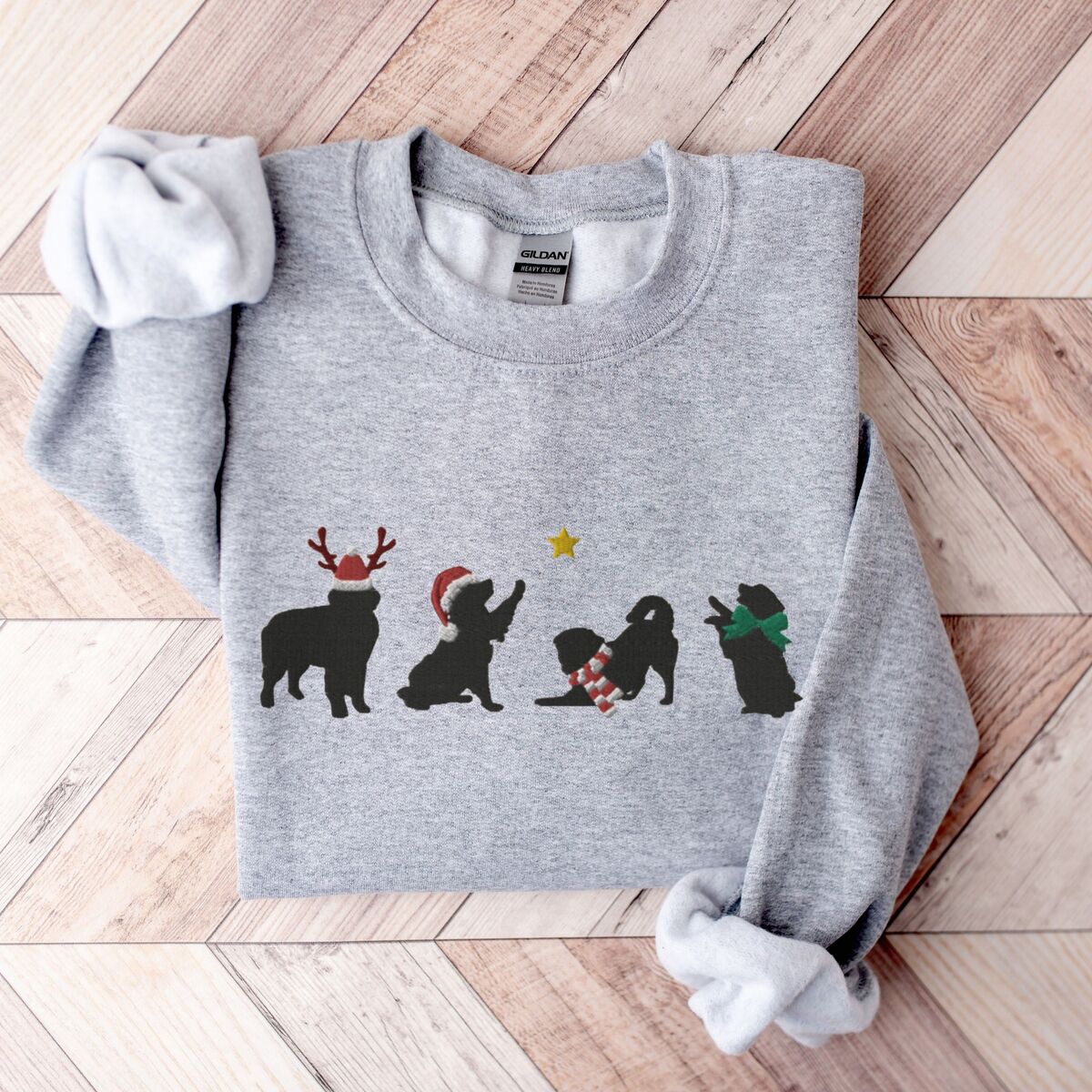 Bernese Mountain Dog Silhouette Sweatshirt - Sport Grey Sport Grey crewneck sweatshirt laid flat on a chevron wood floor, embroidered with four black Bernese mountain dog silhouettes in antlers, a Santa hat, a striped scarf and a green bow under a gold star