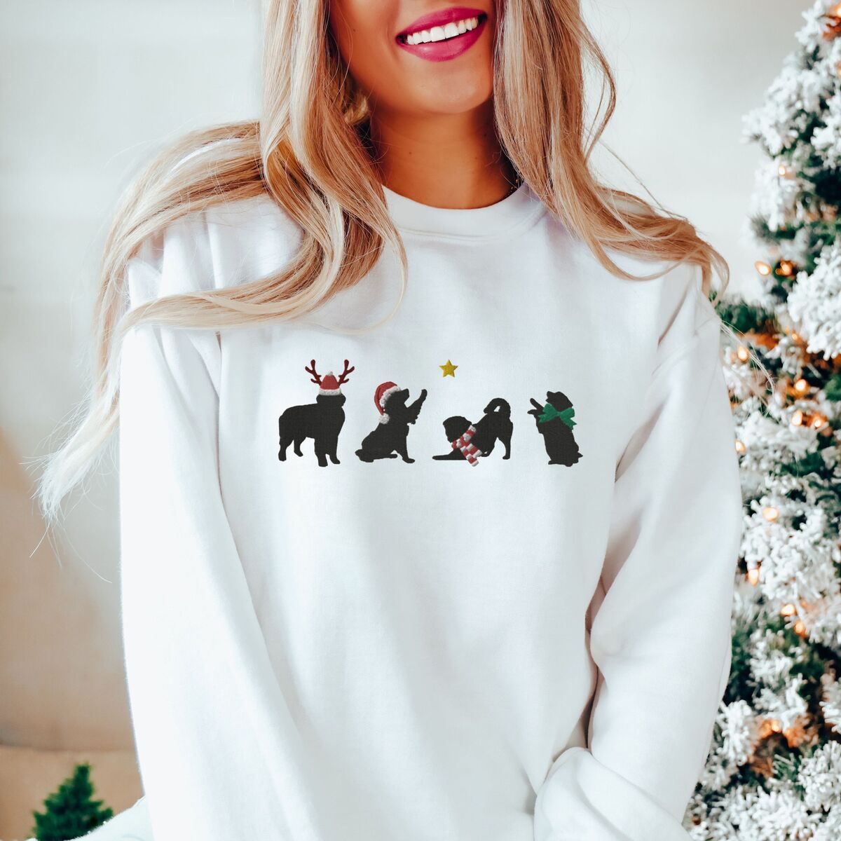 Bernese Mountain Dog Silhouette Sweatshirt - White Smiling woman by a snowy Christmas tree in a White crewneck sweatshirt embroidered with four black Bernese mountain dog silhouettes in antlers, a Santa hat, a striped scarf and a green bow under a gold star