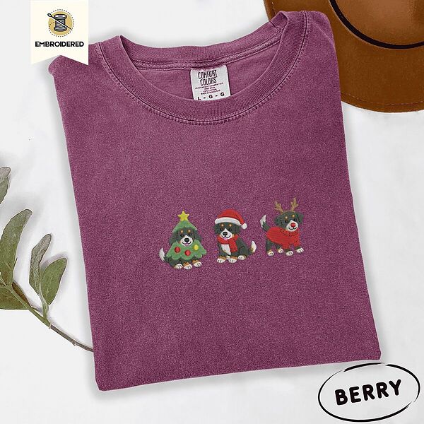 Berry Comfort Colors T-shirt embroidered with three tricolor Bernese mountain dog puppies in a Christmas tree costume, a Santa hat with a red scarf and reindeer antlers with a red sweater