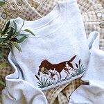 Ash crewneck sweatshirt on gingham overalls with olive branches, embroidered with a liver and white bird dog carrying a mallard duck through cattails and marsh grass
