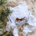 Ash crewneck sweatshirt laid on gingham overalls and a knit blanket with olive branches, embroidered with a bird dog retrieving a mallard through cattails