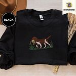 Black crewneck sweatshirt with a brown felt hat and dried bunny tail grass, embroidered with a liver and white bird dog carrying a mallard duck through cattails and marsh grass
