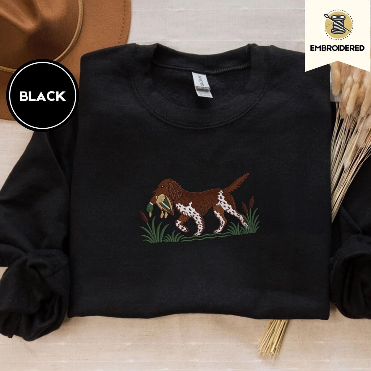 Bird Dog Embroidered Sweatshirt – Black Black crewneck sweatshirt with a brown felt hat and dried bunny tail grass, embroidered with a liver and white bird dog carrying a mallard duck through cattails and marsh grass