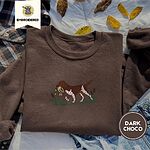 Dark Chocolate crewneck sweatshirt on a plaid flannel shirt with yellow autumn leaves and denim shorts, embroidered with a liver and white bird dog carrying a mallard duck through cattails and marsh grass